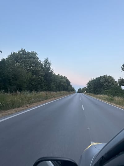 This image depicts a road stretching into the distance, lined by lush green trees on either side. The sky is a soft, clear blue, transitioning to a gentle pinkish hue near the horizon, suggesting it is either early morning or late evening. A faint white line divides the asphalt road, indicating it's a two-way street. In the foreground, the right side of the image shows a portion of a car, including its side mirror and part of the hood, with a yellow reflection on the car's body. The perspective suggests the photo was taken from inside the car, looking forward. The scene appears peaceful and serene, characteristic of a drive through the countryside. The location is identified as Debelets, Bulgaria. There is no visible text in the image.