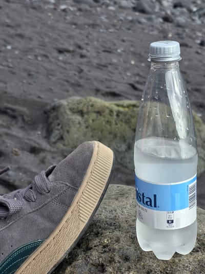 This media file shows a close-up of a partially filled bottle of Kristall mineral water resting on a rough, dark-colored rock. The bottle, made of clear plastic, has condensation on its exterior. The label on the bottle is primarily white with a light blue band that features the brand name "Kristall" in dark blue, along with text indicating "EAU DE SELTZ" and nutritional information. Specifically, it states "250 ml 0 kJ 0 kcal" and "700 ml: 0 kJ/0 kcal." A barcode and the number "2017768" are also visible on the label.

In the foreground, a portion of a grey suede sneaker is visible, placed on a dark, textured surface that appears to be a rocky beach or shoreline. The sneaker has grey laces and a light beige sole with a ribbed texture. The background is blurred, showing a grey, pebbly ground and another large, moss-covered rock. The lighting suggests it might be daytime, possibly overcast, given the diffused light and lack of strong shadows. The overall impression is a casual, outdoor scene, likely in a natural setting.