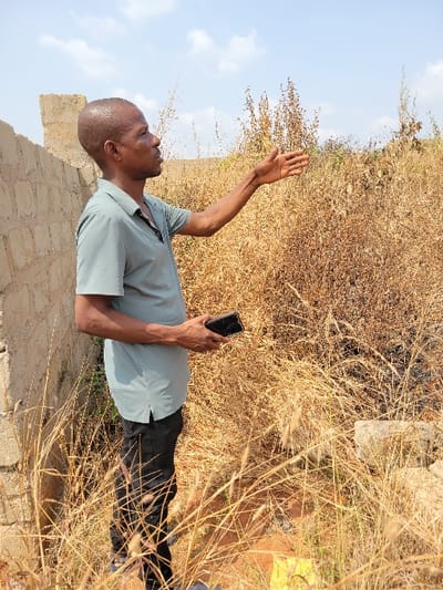 A male subject is depicted standing outdoors in the city of Nwofia, Nigeria. He is positioned in profile, facing right, wearing a light teal short-sleeved collared shirt and dark trousers. His right arm is extended and slightly raised, with the hand open and palm facing right, indicating a gesture. A black smartphone is held in his left hand, positioned near his waist. To the subject's left, a light-colored cinder block wall, appearing partially constructed or in disrepair, is visible. The ground and background are predominantly covered with dense, dry, golden-brown grass and weeds. The upper background features a light blue sky with scattered white clouds. A small, indistinct yellow object is partially visible on the ground in the lower right foreground.