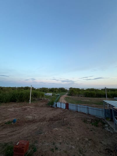 The image shows a landscape in Красный Бор, Russia. The sky, predominantly blue with some light clouds, takes up a significant portion of the left side of the frame. The right side reveals a scene of rural development. A dirt road stretches into the distance, flanked by vegetation. There is also some construction, possibly related to the development. Visible objects include construction materials, a partial structure covered with sheets of corrugated metal, utility poles, and greenery. The ground appears to be recently turned soil. The lighting suggests it is either early morning or late afternoon.