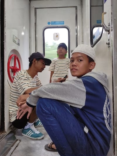 Three young males are captured in a bright, upward-angled shot inside what appears to be a train carriage in Kecamatan Beji, Indonesia. The overall setting is a utilitarian train interior with white walls and a metallic, diamond-plate textured floor.
In the foreground, a young boy with light skin and dark hair, wearing a white crochet kufi (skullcap) and a blue hooded jacket with grey sleeves, crouches and looks directly at the camera with a neutral, slightly serious expression. His forehead glistens, possibly from sweat or light reflection. His hands are clasped between his knees.
Behind him, to the left, a second boy with a darker complexion, wearing a black baseball cap and a light-colored striped t-shirt, is seated on the floor. He leans back against the train wall, looking slightly upwards. His hands rest on his lap, and he wears light blue athletic shoes with white stripes.
Further back and slightly obscured, a third young boy, also in a striped t-shirt and dark cap, stands or leans by a closed train door. He is looking out through the door's window, where bright green outdoor scenery is visible, indicating the train is moving during the daytime.
Notable objects within the carriage include a silver door handle on the white door. Below the door's window, a blue rectangular sign with white text is visible; it features a white symbol of a trash can with a diagonal line through it, implying "No Littering," and partially legible text "DILARANG MEMBUANG SAMPAH SEMBARANGAN" (Forbidden to litter). Near the seated boy, a prominent red handwheel, possibly an emergency brake or valve, is attached to the wall. Above this wheel, a small white rectangular sticker with black, mostly illegible text is present, with faint words like "REDO" and "MAX" discernible. The atmosphere appears casual and relaxed, indicative of travel.