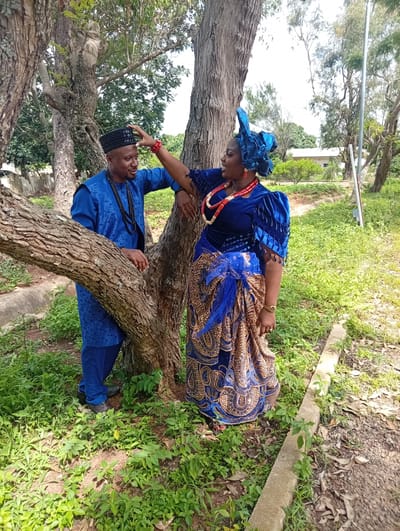 A man and a woman in traditional Nigerian attire pose in an outdoor, grassy area with trees. The man, wearing a blue outfit and a black patterned cap, is leaning against a tree. The woman, also in blue with a matching head wrap and a patterned skirt, stands facing him, her hand touching his head. Both are adorned with jewelry, including red beaded necklaces. The background shows more trees, a building, and a light pole, suggesting a semi-urban or park-like setting. The ground is covered in lush green grass and some fallen leaves. The lighting indicates it is daytime.