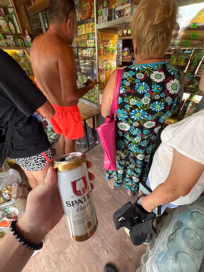 A first-person perspective shot shows a hand holding an open can of Spaten beer. The hand is wearing a black beaded bracelet. In the background, a shirtless man in bright orange shorts stands near a shelf stocked with goods. A woman in a black dress with a floral pattern and a pink bag is also visible, as is another person in a white shirt. The scene appears to be inside a small shop or market, potentially in Sylva, Russia, given the context. The shelves are filled with various packaged food items. The lighting suggests an indoor setting, possibly with natural light coming from the front.