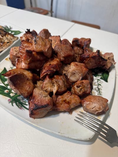 A close-up, high-angle shot shows a white rectangular plate filled with grilled meat cubes. The meat is various shades of brown, with some pieces exhibiting char marks. The plate has a decorative pattern of green leaves and small white flowers along one edge. To the right of the plate, a silver fork rests on the white surface of a table. In the background, the blurry image of a chair and part of a wall can be seen. The setting appears to be a domestic dining area. The location in the city of Бигила, Russia, is unknown.