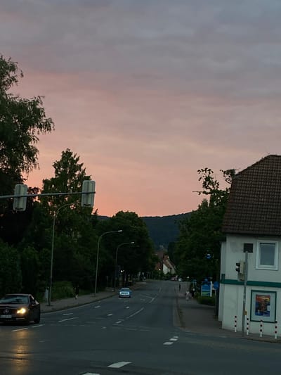 The image depicts a street scene in Rinteln, Germany, during dusk. A street with cars, traffic lights, and pedestrian activity is visible. On the right side of the street, a building with a tiled roof and a store window display is present. Trees line the street, and a hill forms the backdrop. The sky is a gradient of pink and gray hues, suggesting the sun has recently set. The overall atmosphere is calm and quiet.