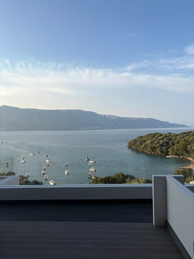 This is a vertical, eye-level, wide shot of a tranquil seascape taken from a balcony in Gouvia, Greece. The foreground is dominated by a dark, textured balcony floor and a light-colored railing. Beyond the railing, the expansive blue water of the Ionian Sea is dotted with numerous small white boats. In the mid-ground, a lush, green, tree-covered island curves into the water, with a small sandy beach and a pier visible. The background features a long, low mountain range under a clear blue sky with wispy white clouds. The overall impression is one of peace and natural beauty.