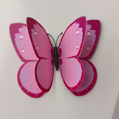 A close-up, eye-level shot shows a pink paper butterfly decoration against a neutral, off-white background. The butterfly has large, intricately patterned wings with lighter pink and silver lines. Several circular, lighter pink spots are visible on the wings. The butterfly's body is black and segmented, with two thin, black antennae extending upwards. The butterfly appears to be attached to the background, likely with adhesive, and is positioned centrally in the frame, with its wings spread open. The lighting is soft and even, creating minimal shadows. There is no discernible environmental context to suggest a specific location within Tohatin, Moldova, or any other city.