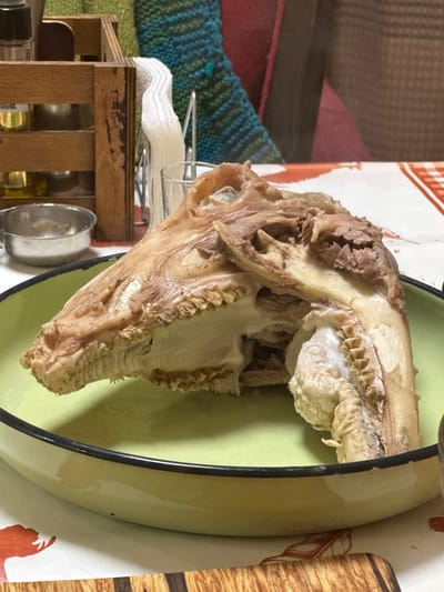 The image shows a sheep's head in a light green bowl. The head appears to have been cooked or boiled. The bowl is sitting on a table covered with a patterned cloth. To the left of the bowl is a wooden crate with spice jars inside. Behind the crate is a dish rack with a stack of napkins. The location of the photograph is purportedly Okop, Bulgaria. No people are visible in the image.