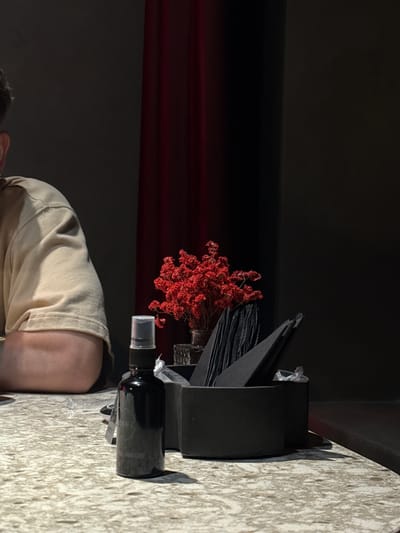 A person is partially visible from the side, wearing a light-colored t-shirt. They are seated at a speckled countertop. In the foreground, a dark spray bottle with a clear cap sits on the counter. Behind the bottle, a black rectangular container holds several black folded napkins. To the left of the napkins, a small glass vase contains a sprig of dried red flowers. The background consists of a dark wall and a deep red curtain. The scene appears to be indoors.