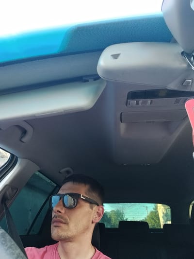 This is a selfie taken from inside a car, looking towards the windshield and the interior roof. A man wearing sunglasses and a pink t-shirt is visible in the lower right of the frame, looking to his left. The car's interior includes the rearview mirror, sun visors, and overhead console. The background shows a blurred view of trees and buildings through the tinted rear window, suggesting a sunny day. The setting is likely a street in Șoldănești, Moldova, given the provided context.