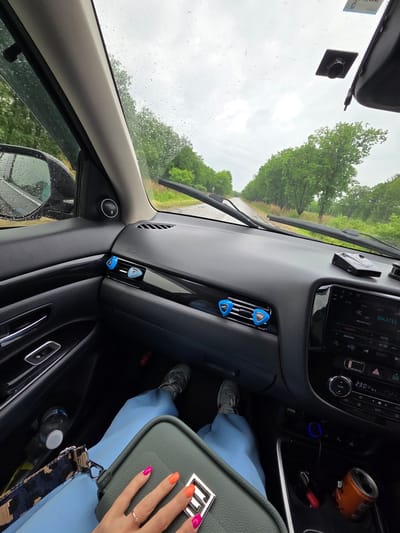 The image captures the interior of a vehicle from a front passenger's perspective, looking forward through a rain-splattered windshield onto an asphalt road. The scene is set in the city of Valdgeym, Russia, under an overcast sky. The road is flanked by dense green trees and recedes into the distance.

Inside the car, the dark-toned dashboard features a glossy black decorative strip across its width. Three blue, guitar-pick-shaped air fresheners are clipped to the air vents—two centrally and one on the far right. A black electronic device, likely a radar detector, is positioned on the dashboard to the right of the center. The infotainment system display shows "ISKATEĽ" (Seeker) and "Музыка рок" (Rock music), along with radio station presets like "DACHA" and "AVTO." The temperature is displayed as "23.0 °C." A blue-lit charging port is located beneath the infotainment system. A JBL brand speaker is visible on the driver's side A-pillar. A rectangular black mount is affixed to the windshield below the rearview mirror, and a partial sticker is visible on the upper right side of the windshield.

The passenger's lower body is visible, clad in light blue trousers and grey sneakers. A clear plastic water bottle with a white cap rests in the passenger footwell, where a red object is also partially seen. An orange beverage can is placed in a cup holder in the center console. The passenger's left hand, adorned with a gold ring on the ring finger and fingernails painted in alternating pink and orange hues, rests on a structured dark green handbag. The handbag features a prominent silver clasp and a patterned strap in shades of brown and black.