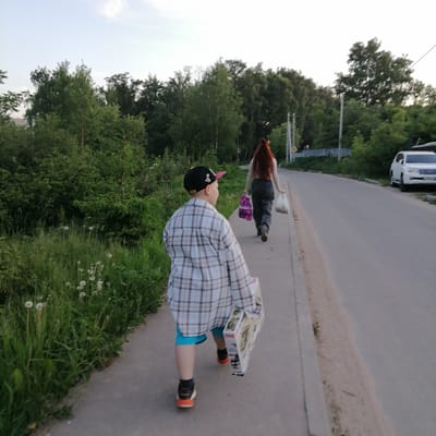 The image captures a candid, rear-view perspective of two individuals, an adult and a child, walking away from the camera on a paved sidewalk in a semi-urban or suburban setting. The time of day appears to be late afternoon or early evening, indicated by soft, diffused natural light and a light blue sky with scattered clouds.

In the foreground, a child, appearing to be a boy, is seen from behind. He wears a black baseball cap with a pink brim, a loose-fitting plaid long-sleeved shirt primarily in white, grey, and brown tones, blue shorts, and black and orange athletic shoes. He is carrying a large, rectangular white box with green graphics and red trim in his right hand.

Ahead of the child, an adult, consistent with a woman, also walks away from the camera. She has long, wavy reddish-brown hair, wears a light-colored long-sleeved top, dark pants, and dark footwear. She carries a purple-patterned shopping bag in her left hand and a white plastic shopping bag in her right hand.

The concrete sidewalk runs parallel to an asphalt road on the right side of the frame. A narrow strip of dirt and short grass separates the sidewalk from the road. The left side of the image is dominated by dense green vegetation, including tall trees and bushes, which form a thick backdrop. Scattered white dandelions and tall grass grow along the edge of the sidewalk. Along the road, several utility poles with overhead wires are visible. A light-colored fence and a small yellow square sign mounted on a pole are discernible in the middle distance on the right. A white SUV is parked facing away from the camera on the right shoulder of the asphalt road in the background.

The specific location in the city of Kryukovo, Russia, cannot be confirmed solely based on the visual evidence within the image.