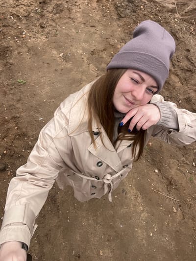 A woman with brown hair and blue nails stands outside, winking and smiling at the camera. She wears a gray beanie and a beige trench coat. The ground is dirt. The location is Ivlevo, Russia.