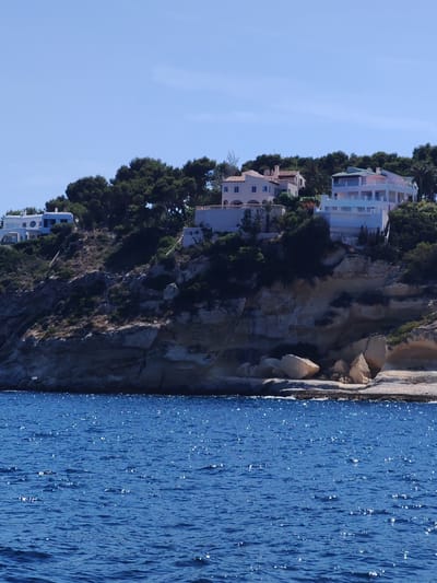 This media file depicts a scenic view of the Mediterranean Sea and a coastal landscape in Sol de Mallorca, Spain. The upper portion of the image showcases several modern, multi-story houses nestled atop a lush green, tree-covered hillside. These residences feature contemporary architectural designs with clean lines, balconies, and large windows, suggesting a luxurious setting. The buildings are varied in color and style, with some exhibiting pale pink, white, and light blue facades. The hillside itself is rocky and uneven, with abundant vegetation, including pine trees and other Mediterranean flora.

Below the hillside, the sea dominates the lower part of the image. The water is a vibrant, deep blue, with a gentle ripple and numerous sparkling highlights from the sun, indicating a bright, sunny day. The sun's reflection on the water suggests that the time of day is likely midday or early afternoon. The weather appears clear and pleasant, with a bright blue sky visible above the houses and trees.

The scene evokes a sense of tranquility and leisure, characteristic of a coastal vacation or residential area. The combination of the beautiful blue sea, the rocky coastline, and the well-appointed houses creates a picturesque and inviting atmosphere. There are no visible people or specific activities taking place, but the presence of the houses implies habitation. The location cues, such as the type of vegetation and the architectural style of the houses, are consistent with a Mediterranean coastal environment. No visible text is present in the image.
