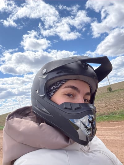 A person wearing a black motocross helmet is seen in a close-up, angled view. The helmet has a visor and a peak, and a bandana is visible around the person's neck inside the helmet. The person's eye is visible. The background is a field and a partly cloudy blue sky. The location cannot be determined to be Mozhga, Russia from the image alone.