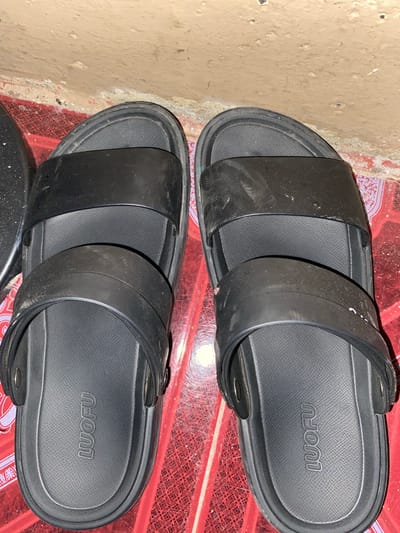 A pair of black, two-strap slides are positioned side-by-side on a red, patterned tiled floor. The slides have a matte finish and a raised textured footbed with the embossed brand name "LUOFU" visible on each insole. The wall behind the slides is a rough, textured, tan-colored surface that appears to be concrete or plaster. The scene is indoors, suggesting a domestic setting or a shop. The lighting is somewhat dim, with a bright light source coming from the right, casting subtle shadows. There are no people present in the image. This scene is located in Lambaro Kueh, Indonesia.