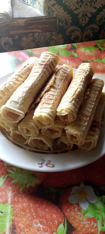 A close-up, eye-level shot shows a white plate filled with rolled wafer cookies on a tablecloth patterned with strawberries. The cookies are golden brown and have a ridged texture. The plate has a subtle floral design around its rim. The background is blurred, but appears to be a room with patterned curtains and a wooden surface. The image is well-lit, suggesting it was taken indoors during the day. No people are visible in the image.