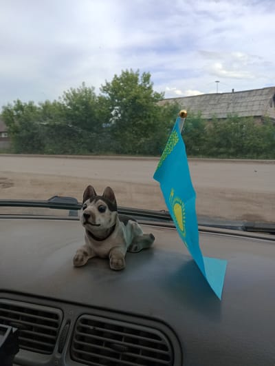 The image depicts a close-up view from inside a vehicle, looking out through the windshield. The dashboard is visible in the foreground, featuring a toy husky dog and a small flag of Kazakhstan. The flag, predominantly blue with a yellow sun and eagle design, is mounted on a small pole with a golden finial. Outside the vehicle, a dusty road runs parallel to a line of green trees. In the background, a weathered wooden building with a gray roof is visible, situated amongst more trees. The sky is overcast with scattered clouds. The location is identified as Atbasar, Kazakhstan. There are no people or specific actions depicted.