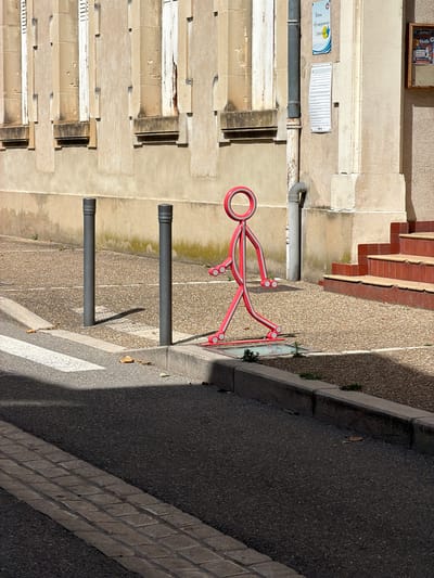 This is an eye-level, outdoor, daytime, medium shot of a street corner with a crosswalk in Mercurol-Veaunes, France. In the foreground, a dark asphalt road runs diagonally from the lower left to the upper right. A white crosswalk is visible on the left side of the road, partially in shadow. A textured gray curb separates the road from a pebbled sidewalk.

On the sidewalk, two dark gray cylindrical bollards stand vertically. To the right of the bollards is a bright pink, stylized figure resembling a person made of metal tubing. The figure has a circular head with a smaller circle inside, and its limbs are articulated. It appears to be in a walking pose.

In the background, a building with a beige stone facade features tall, narrow windows with white shutters. A gray downspout runs vertically along the side of the building. To the far right, a set of red tiled stairs ascends. A small sign with blue and white text is attached to the building near the downspout.