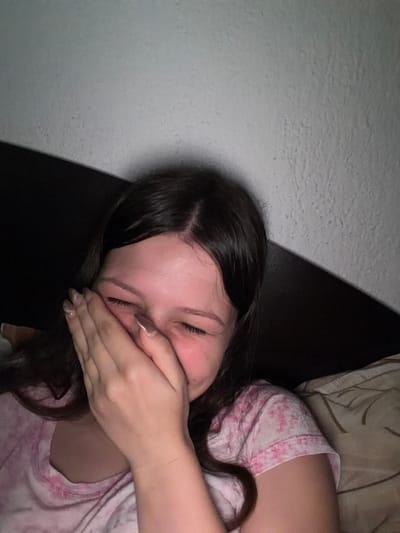 A young person with dark hair and fair skin is lying down, covering their mouth with their hand. The person appears to be on a dark-colored surface, possibly a bed or couch, next to a white wall. The person is wearing a pink and white patterned top and there is a pillow behind them. The location is not identifiable as Parvomai, Bulgaria based on the image alone.