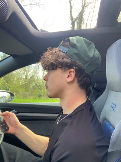A male individual is observed in profile, occupying the driver's seat of a vehicle. He has dark, curly hair, styled with faded sides and a longer back, and wears a dark green baseball cap backward. A silver chain necklace is visible around his neck, which also bears a reddish mark on the left side. He is dressed in a black t-shirt. His right hand rests on the vehicle's steering wheel, while his left hand holds a silver-toned smartphone displaying an illuminated music player interface. The vehicle's interior features black trim, and a dark grey/blue sport seat with a blue "R" emblem is visible on the passenger side. Through the windows and sunroof, green foliage, trees, and a bright sky are apparent, indicating an outdoor, possibly semi-rural environment. A blurred reflection in the driver's side mirror suggests the presence of distant buildings or structures. The scene is located in Broxbourne, United Kingdom.