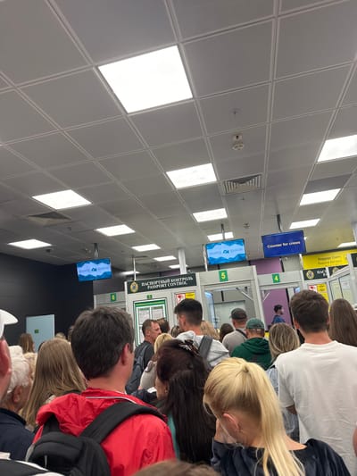 This image shows a crowded line of people in what appears to be an airport, based on the signs visible in the image. Signs in Russian are visible as well as English. "Counter for crews" is one of the signs. The area features a tiled ceiling with fluorescent lights. People are mostly obscured, with only the backs of their heads and some of their clothing visible.