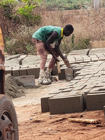 The scene depicts an outdoor brick-making or construction site in Nwofia, Nigeria. An adult male with a dark complexion, wearing a sleeveless green and yellow patterned shirt, reddish-brown trousers, and light-colored fabric wrapped around his lower legs, is positioned in a bent posture over rows of concrete blocks. He is actively engaged, with his hands near a textured metal brick mold or tool. Numerous gray rectangular concrete blocks are arranged in neat rows on the reddish-brown earth, presumably drying. To the left, a pile of light gray, sandy material is visible. In the lower left foreground, a section of a large, dark vehicle tire with a rusty metal hub is present. A long, pale object, possibly a root or piece of wood, lies on the ground in the lower right foreground. The background features dry, sparse vegetation and some green foliage under an unidentifiable sky.