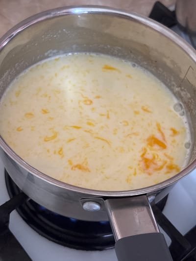 A close-up, overhead view of a stainless steel saucepan filled with a creamy, pale yellow liquid containing small, irregular pieces of bright orange, possibly citrus zest. The saucepan is placed on a gas stovetop, with a faint blue flame visible beneath the burner. The liquid appears to be gently simmering, with small bubbles on the surface. The handle of the saucepan, made of black plastic and metal, is visible in the foreground. The background is blurred but suggests a domestic kitchen environment with tiled countertops. There are no people or animals present. This scene is located in the city of Новый Кинер, Russia.