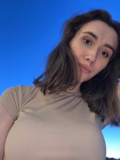 A close-up, low-angle selfie shows a woman against a clear blue sky. The woman has medium-length brown hair and is wearing a tan, ribbed t-shirt. Her face is angled towards the camera, with her lips slightly parted. The background is an uninterrupted expanse of blue sky. There are no visible buildings, structures, or natural landmarks to indicate a specific location, including Sharovechka, Ukraine.