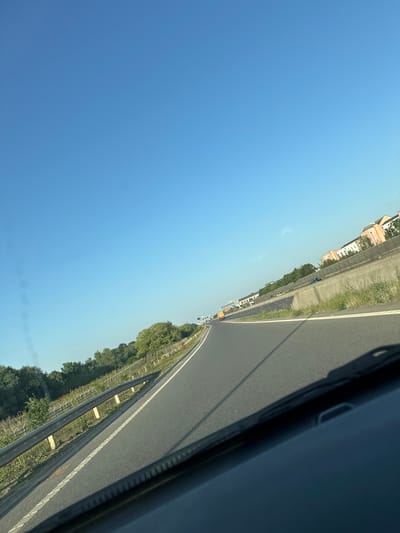 This media file depicts a view from a vehicle on a highway, likely in South Cambridgeshire, United Kingdom. The scene is captured from a slightly tilted perspective, looking down a road that stretches into the distance. The sky is a clear, bright blue, suggesting a sunny day.
On the left side of the road, a line of buildings with light-colored facades are visible, interspersed with trees. Further down the road, more buildings and a white truck can be seen. The right side of the road features a grassy verge and trees, with a metal guardrail separating the road from the verge. The asphalt of the highway is in good condition, with white lines marking the lanes. A dark shadow, possibly cast by the vehicle or a passing object, is visible on the right side of the frame, extending across the road.
The activity appears to be a journey along the highway, with other vehicles present in the distance. The time of day is likely midday, given the bright sunlight and clear sky. There are no discernible emotions conveyed, and no visible text in the image. The overall impression is one of a typical daytime drive on a highway in a rural or semi-rural setting.