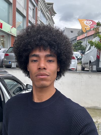 A man with dark, curly hair and a navy blue sweater takes a selfie on a street in Güngören, Turkey. A car door is open to the right of the man, with another grey car parked behind it. Across the street is a building with a "MANCHE" sign. A Galatasaray flag is visible. The sky is overcast.