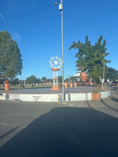 In the city of Bulukumba, Indonesia, a monument with a ship's wheel as its centerpiece stands in a circular plaza. The monument is white with orange accents and is surrounded by trees and other greenery. The sky is a clear blue, and the sun is shining. The plaza is paved, and there are a few people walking around. The area appears to be a public space.