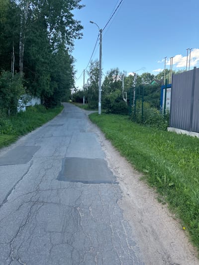 The image shows a moderately cracked asphalt road with several patches, suggesting it is in a state of disrepair. The road is lined with lush green grass and trees on both sides. On the right side of the road, there is a metal fence and a building with signage that appears to be in Russian. The sky is clear with a few white clouds. The scene appears to be a quiet street in a rural or suburban area. Based on the signage, the location is likely Yanino-2, Russia.