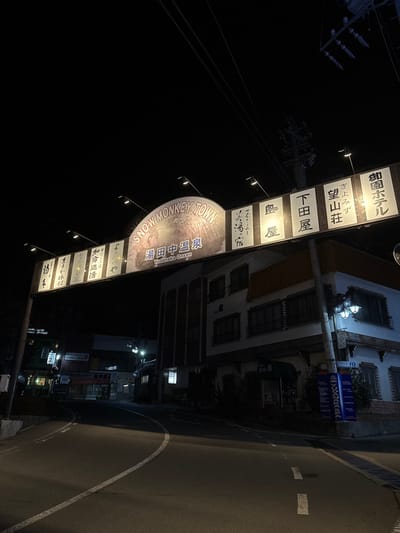 The image displays a nighttime street scene in Yudanaka Onsen, Yamanouchi, Japan. An illuminated arch-style sign spans across an asphalt road, prominently featuring the text "SNOW MONKEY TOWN" and "湯田中温泉 Yudanaka Onsen" on its central panel, which also includes an illustration of a snow monkey. Flanking this central section are multiple rectangular panels with Japanese script, identifying local businesses or accommodations such as "御園ホテル" (Misono Hotel) and "下田屋" (Shimotaya).

The road, marked with white lines, curves slightly to the left and is clear of traffic and pedestrians. On the immediate right, a two-story building features a light-toned lower facade, a darker upper level, decorative window surrounds, and a wall-mounted streetlamp. A vertical blue banner sign on the building's corner displays "YUDANAKA SPA" and other Japanese text. Distant buildings are partially visible and dimly lit along the street. Overhead power lines and utility poles are discernible against the dark, clear night sky.