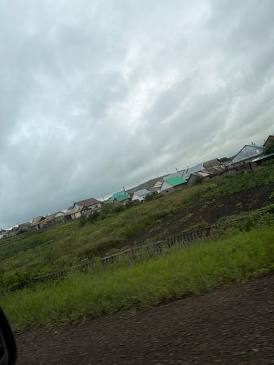 The image shows a street in Kendektamak, Russia, with a row of houses on the left and a field on the right. The houses are small and appear to be residential. The field is covered in grass and some dirt patches. A dark, textured surface, possibly a road or a path, occupies the right side of the image. The sky is overcast with gray clouds, suggesting a cloudy or possibly rainy day. The overall scene depicts a rural or semi-rural setting in a Russian village. No people or specific activities are clearly visible. The orientation of the image is sideways, making it appear as if the scene is tilted.