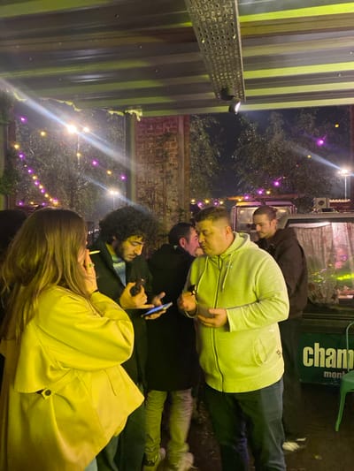A group of people gather outdoors at night. String lights illuminate the scene. One man in a black coat holds a phone. Another man wears a light green hoodie. To their right, a man in a dark brown coat looks downwards. A woman in a yellow jacket is in the foreground, seemingly smoking. The background includes a metallic structure and greenery.
