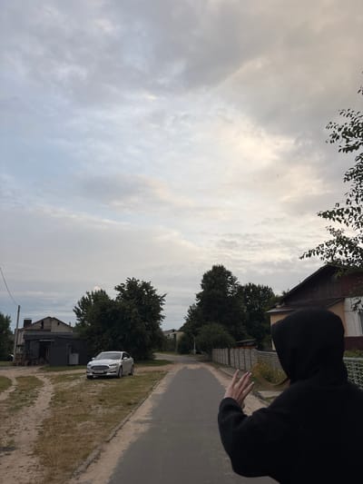 The image depicts a street scene in a rural area. A person wearing a black hoodie with the hood up is standing on a paved road, facing away from the camera and towards a grey sedan parked on a dirt and grass area to the left. The person's right hand is raised, with fingers spread, as if waving or posing for a photo. Behind the car is a low, dark-colored building, possibly a shed or garage, with some wooden pallets stacked nearby. Further in the background, there are trees and houses. The sky is overcast with a mix of grey and lighter clouds. The overall setting suggests a quiet, possibly residential, area in the countryside. The car's license plate is visible, with markings that may be consistent with Belarusian plates.