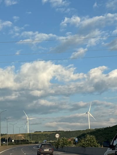 This image depicts a highway scene in Otarsheni, Georgia, under a partly cloudy blue sky. Two wind turbines stand prominently on a distant green hillside. The foreground features a multi-lane highway with several vehicles, including a dark-colored SUV in the right lane. Streetlights line the left side of the road. A speed limit sign indicating 110 is visible on the right. The environment is hilly and rural, with trees and vegetation dotting the landscape.