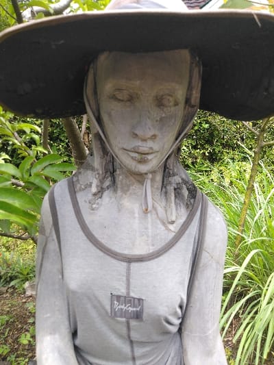 A weathered, light grey concrete or stone sculpture of a female figure is positioned outdoors in Ba Nwet Kone, Myanmar. The sculpture, seen from the chest up, wears a dark, wide-brimmed hat and a light grey long-sleeved top with a dark grey trim around the neckline and a vertical seam down the center front. A rectangular patch displaying the word "BodyGuard" in a dark cursive-style font is affixed to the upper left chest. The figure's face features closed eyes, a straight nose, and closed lips, with a light grey hood-like element framing the face and extending down the neck. The background consists of vibrant green foliage, including broad leaves on the left and tall, slender leaves on the right, under natural daylight.