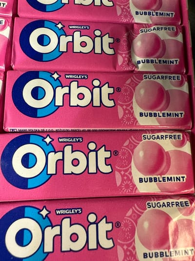 A close-up photograph displays a partial row of "Orbit" bubblegum packets. The packets are primarily pink and feature a large "Orbit" logo in white and blue. Above the logo, the word "WRIGLEY'S" is printed in a smaller font. At the top of each packet, the flavor is indicated: "BUBBLEMINT" and "SUGARFREE" alternate. The packets are lined up next to each other. There is no indication of a location in Darien, United States of America.