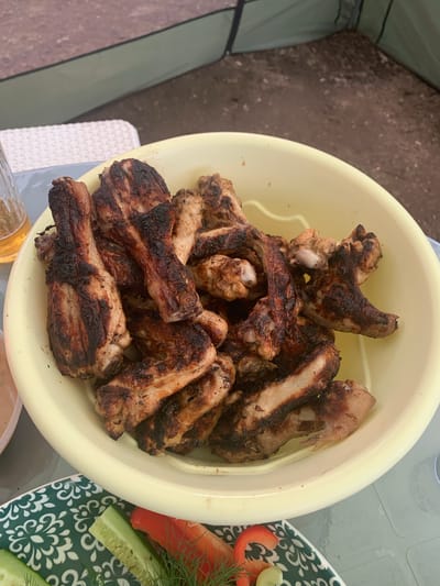 A close-up, eye-level shot shows a large, pale yellow bowl overflowing with grilled chicken wings and drumettes. The chicken pieces are visibly charred in places, indicating they were cooked over an open flame, likely on a barbecue. The scene appears to be outdoors, with hints of a tent or canopy in the background, suggesting a picnic or camping setting. A glass of amber liquid, possibly beer, is visible to the left of the bowl. In the foreground, a plate with sliced cucumbers, red bell pepper, and dill is partially visible. The overall impression is of a casual, outdoor meal. The image was taken in Krestzy, Russia, suggesting a local gathering or event.