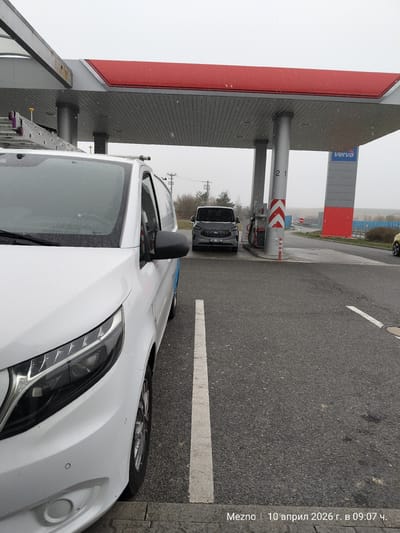 The image depicts a gray van parked at a gas station in Mitrovice, Czechia. Another white van is parked to the side. The gas station has a red and gray canopy with a "verva" sign. The time and date stamp on the photo indicates "Mezno | 10 April 2026 at 09:07". It is a cloudy day with what appears to be a light rain or snow.