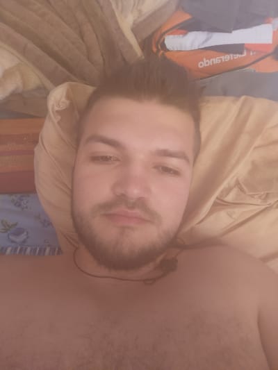The media file is a close-up, over-the-shoulder selfie of a young man lying in bed. The man has short, dark hair styled upwards, a beard, and is looking directly at the camera with a neutral expression. He is partially undressed, with his chest visible. He is wearing a thin dark cord around his neck, possibly from earbuds or a necklace.
The setting appears to be a bedroom. The background shows a rumpled beige pillow and bedding. To the left of the pillow, a striped wooden surface is visible, possibly the headboard of a bed or a shelf. Further to the left and lower down, a blue fabric with a floral pattern is visible, suggesting decorative bedding or a blanket. Behind the man's head and to the right, an orange bag with white and black accents is partially visible. White text on the orange bag appears to read "obnsiets."
The lighting suggests it might be daytime, but there are no clear indicators of the weather or time of day. The overall mood is relaxed and informal, typical of a personal selfie. The location context provided indicates the image was taken in Grigoriopol, Moldova.