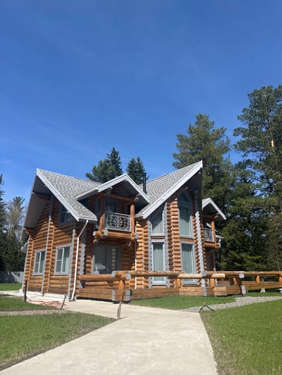 A medium shot of a large log house. The house has a grey shingled roof and is surrounded by green grass and trees. The sky is a clear blue. The location is Areyskoye, Russia. There are no people visible.