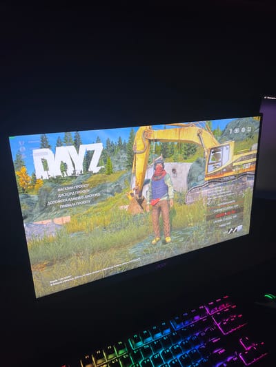 The image shows a computer monitor displaying the game "DayZ". The game's main menu is visible, featuring a character standing in front of a yellow excavator. Text in Russian and Ukrainian is present on the screen, indicating game options and server information. The monitor is placed on a desk with a colorful, backlit keyboard in the foreground. The background is dark, suggesting the image was taken in a dimly lit room. There is no visible indication of the city of Skawina, Poland, or any specific location within it.
