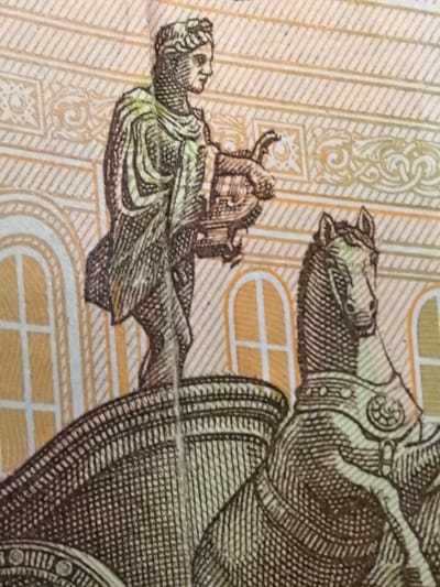 The image is a close-up of a banknote featuring an engraved illustration. In the foreground, a chariot is depicted, drawn by a horse that is mostly visible. The horse is adorned with decorative armor, including a patterned breastplate. A figure stands in the chariot, holding a lyre. This figure is depicted in profile, wearing a draped garment and a headdress. The background of the banknote features architectural elements, including arched windows with cross-shaped panes, and decorative patterns of lines and swirls. The location in the city of Ново-Талицы, Russia is not visible in this image.