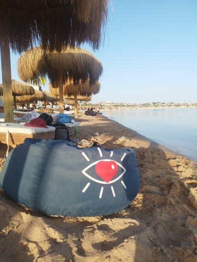 The image depicts a serene beach scene at Ras Abu Soma, Egypt, under a clear, bright blue sky. In the immediate foreground, a dark blue inflatable beanbag, adorned with a distinctive white graphic of an eye featuring a red heart as its pupil, rests on the golden-brown sand, casting soft shadows. The textured sand shows minor undulations and imprints.
Further back on the left, several traditional thatched straw umbrellas are scattered across the beach, offering shade. Beneath them are comfortable white sunbeds, some occupied by individuals relaxing. Personal belongings such as backpacks (one blue, one black), a red hat, and articles of clothing are placed on or near the sunbeds, indicating a leisurely stay. People are visible lounging on the beds or strolling along the shore.
To the right, the calm, inviting turquoise sea gently laps against the sandy beach. The water stretches into the distance, reflecting the clear sky. Along the far horizon, a series of modern resort buildings and structures line the coastline, suggesting a vibrant tourist destination. More distant figures can be seen enjoying the water and the beach.
The lighting indicates either a pleasant morning or late afternoon, characterized by soft sunshine and elongated shadows. The overall atmosphere is one of tranquility and vacation, with perfect weather for beach activities. No explicit textual information is present, only the stylized eye graphic on the beanbag.