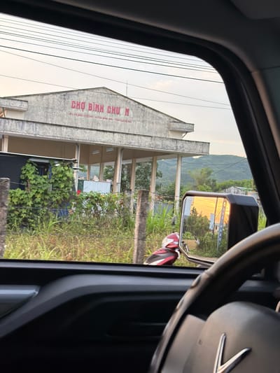 This media file captures an exterior scene in Bình Chương, Vietnam, viewed from the interior of a vehicle. The image is oriented vertically but depicts a landscape view from a car window.
On the left side of the frame, outside the vehicle, a concrete building with a prominent gable roof is visible. Large red text on the building clearly spells out "CHỢ BÌNH CHƯƠNG," which translates to "Bình Chương Market." Below this, smaller text further specifies: "ĐỊA ĐIỂM KINH DOANH CHỢ HỘ - XÃ BÌNH CHƯƠNG - HUYỆN BÌNH SƠN - TỈNH QUẢNG NGÃI," identifying it as a market business location within Bình Chương commune, Bình Sơn district, Quảng Ngãi province. Utility lines crisscross the sky above the building. Green foliage, including bushes and grass, grows in front of and around the building. In the background, rolling green hills or mountains are visible under a pale, soft sky.
To the right, elements of the vehicle's interior are seen, including the dark frame of the car window and a portion of a dark steering wheel. A side-view mirror captures a reflection of warm, golden light, possibly from a setting or rising sun, illuminating the road or landscape behind the vehicle. Partially visible below the mirror is a red and white object, possibly part of a scooter or motorcycle. The overall lighting suggests either early morning or late afternoon. The scene conveys a moment of observation while driving through or stopping near a local market in a rural or semi-urban Vietnamese setting.