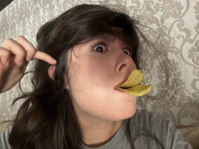 The image is a close-up, medium shot of a young woman with dark hair, sitting in what appears to be an indoor setting. She is looking directly at the camera with wide, surprised eyes and has two potato chips placed on her lips, giving the impression that she is trying to hold them there. Her left hand is raised, with her index finger touching her ear, as if in a gesture of innocence or perhaps to steady herself. The background is a textured wallpaper with a floral pattern in muted tones of beige and grey. The lighting is somewhat dim, suggesting it might be evening or a poorly lit room. The woman is wearing a plain grey t-shirt. The overall mood conveyed is playful and humorous, with the woman's expression and the unusual placement of the chips contributing to this. There is no visible text in the image.