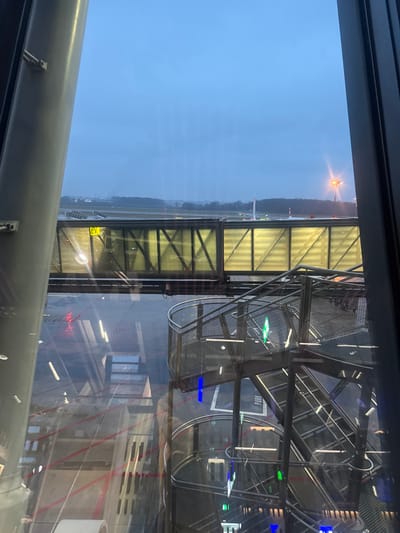 The image captures an airport scene from an elevated perspective, likely from inside a terminal. A jet bridge extends towards an aircraft visible in the distance on the runway. The sky is overcast. The jet bridge is metallic, with transparent panels offering a view of the tarmac. Reflected in the glass, architectural elements of the terminal are visible, creating a layered and slightly abstract composition. Metal stairs surround the jet bridge.